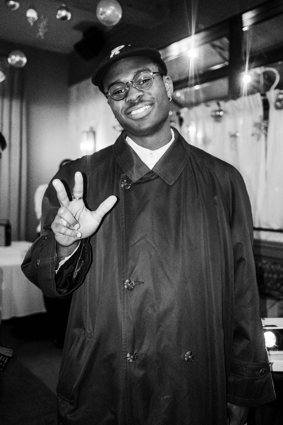 guest in a long trench coat and cap signing to the camera