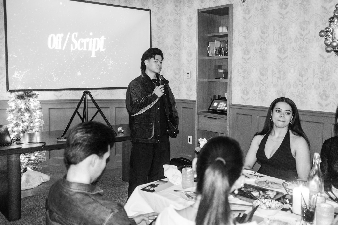 the host on the mic in front of a projector screen reading off/script